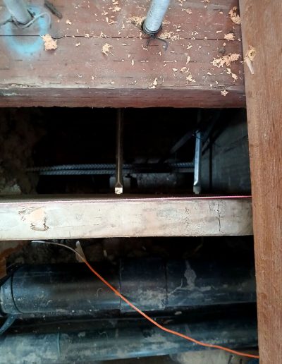 Close-up view through wooden floor framing showing existing plumbing pipes and a rebar element beneath a structure being renovated.