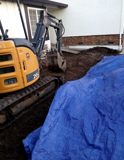 Excavator machinery prepares the ground for a concrete foundation addition near the side of a house.