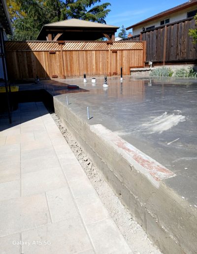 Freshly poured and cured concrete slab foundation with exposed stem wall next to existing patio pavers.