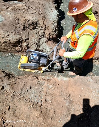 Construction worker in high-visibility vest compacting the gravel base layer in a foundation trench with a plate tamper.