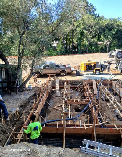 Aerial view of a large residential foundation site with complex formwork, workers, and heavy machinery, including a concrete pump.