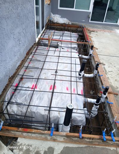 Overlooking a narrow concrete slab area with rebar, vapor barrier, and pre-installed plumbing pipes for a home addition foundation.