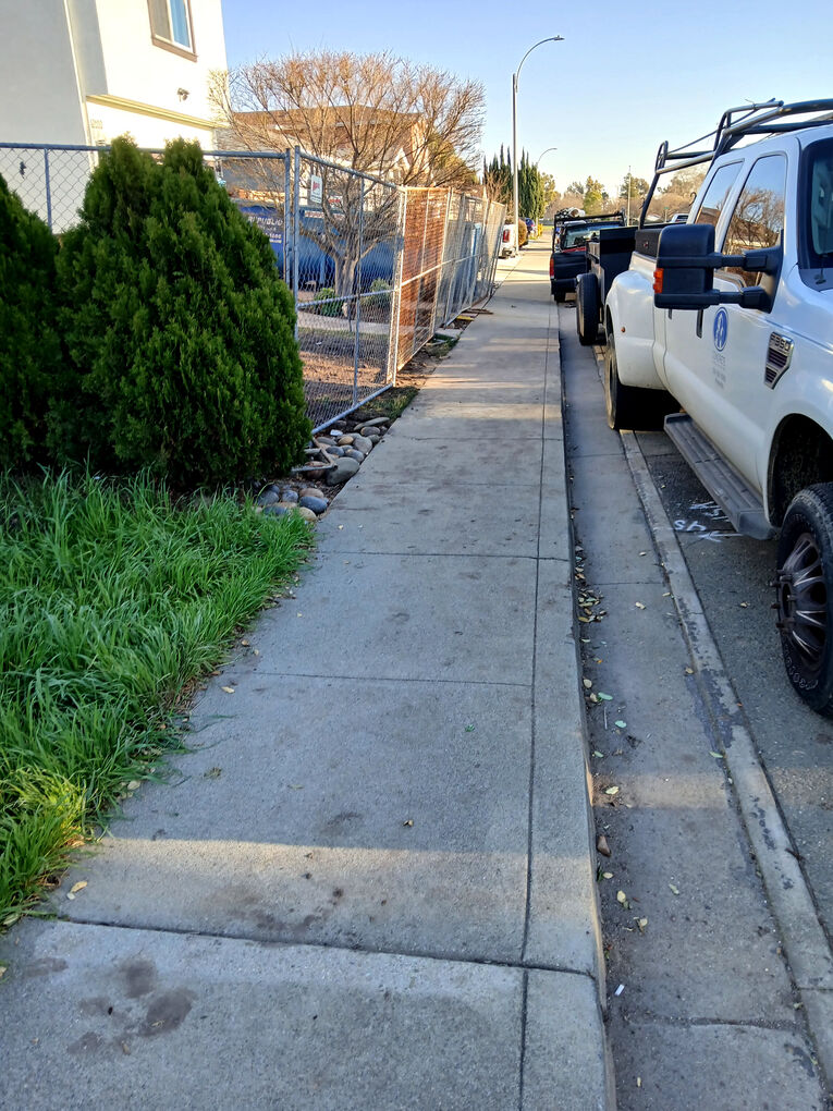 Street-level view of a sidewalk and curb near the work site, showing site fencing and a parked work truck.