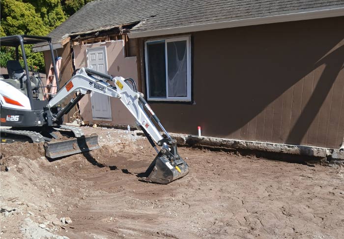 White Bobcat excavator performing heavy-duty site grading and soil removal next to a house wall to prepare the ground for a new concrete foundation or addition.