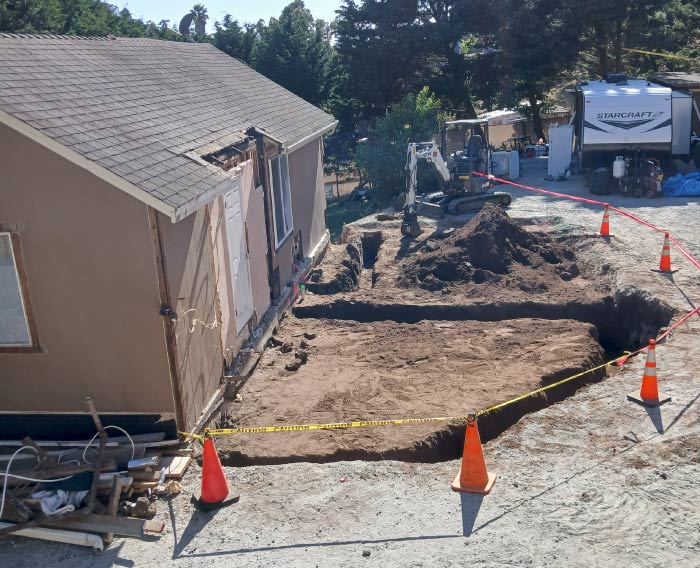 Residential construction site featuring a white Bobcat excavator digging trenches for a driveway expansion, secured with yellow caution tape and orange cones during heavy-duty site preparation