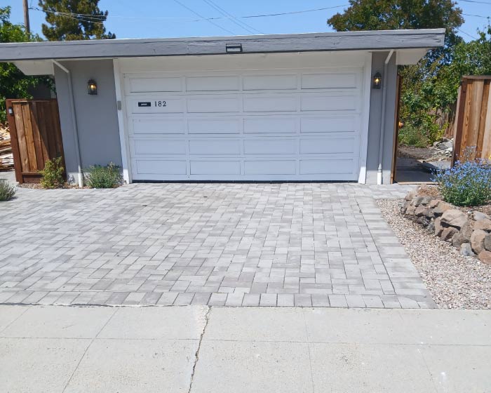 Gray interlocking driveway pavers installed in a classic herringbone pattern leading to a white garage door, often compared against stamped concrete for durability and cost.