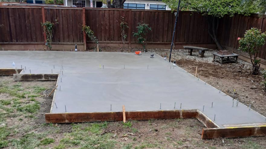 Newly poured residential concrete foundation slab with wooden formwork and embedded steel anchor bolts ready for framing in a backyard.