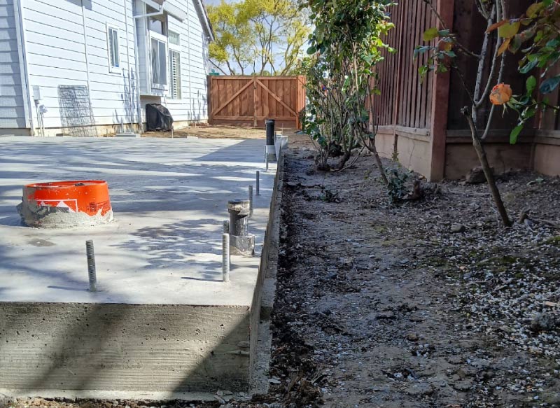 Close-up of a thick, newly finished concrete slab-on-grade foundation showing exposed metal anchor bolts and black PVC plumbing pipes next to a home.