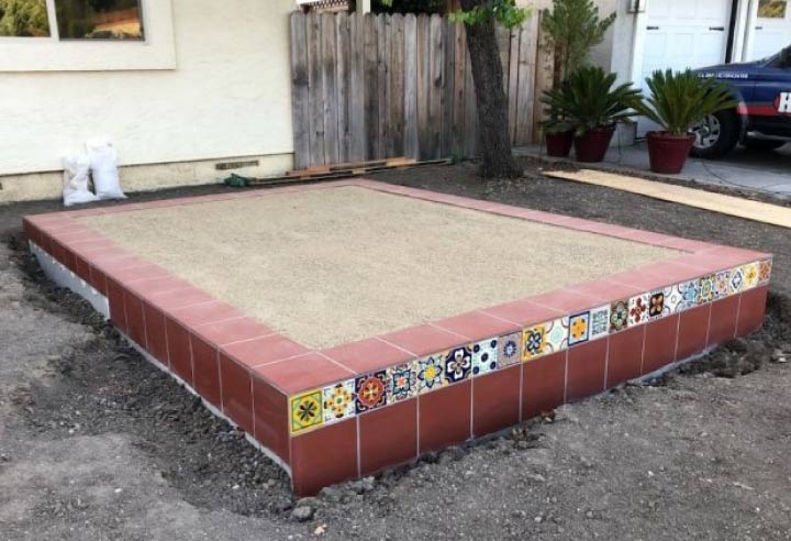 Raised backyard patio or structural base featuring a decorative red and patterned Spanish tile border with a sand-filled interior.