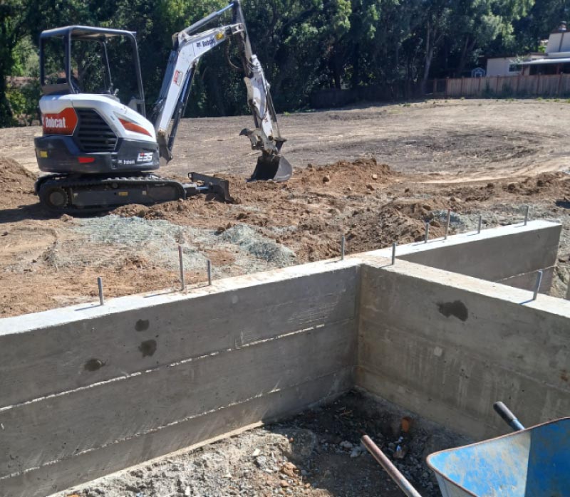 A Bobcat E35 compact excavator performing site preparation and earthmoving next to a newly poured, high-quality concrete retaining wall with integrated anchor bolts for an ADU project.